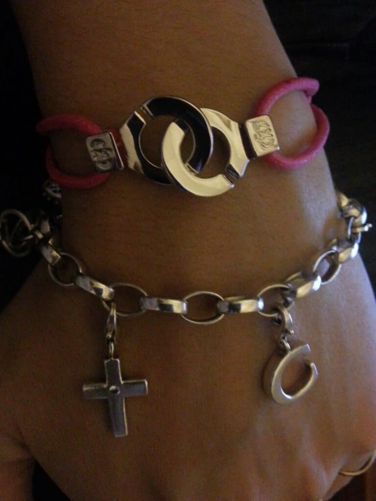 A hand wearing two bracelets: one is a pink cord bracelet with interlocking silver and black rings; the other is a silver chain bracelet with a dangling cross charm and a small silver horseshoe charm. The skin tone is light brown.