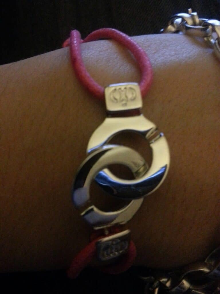 A close-up of a wrist wearing a bracelet with two interlocking silver rings and bright pink cord. The rings are smooth and shiny, and small square silver accents with an engraved logo hold the cords to the rings. Part of another chain bracelet is visible.