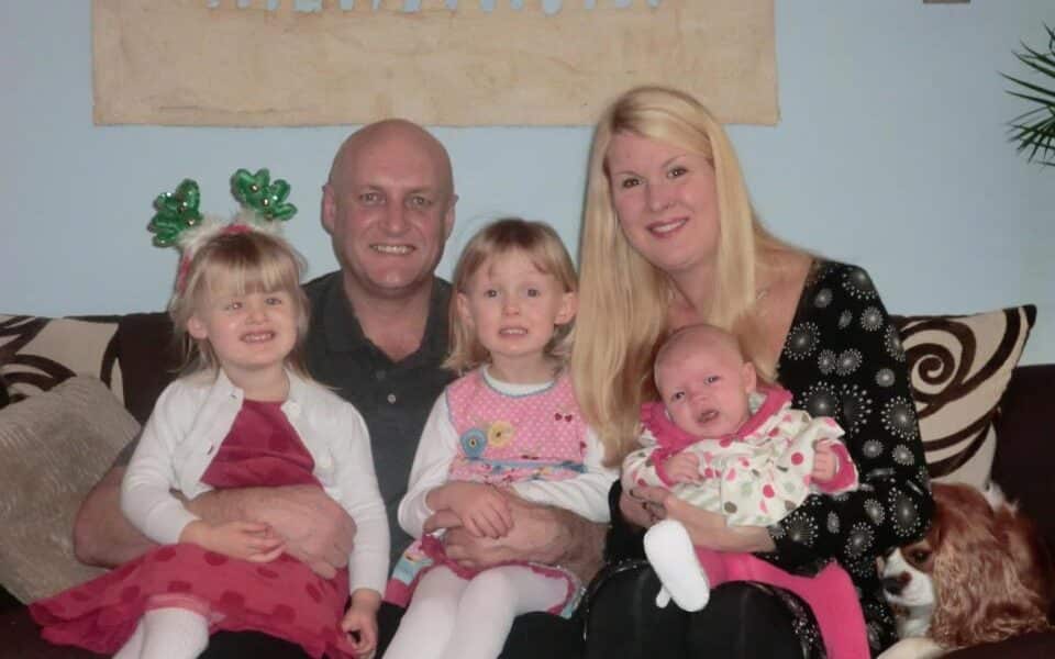 A family of five sits closely on a brown sofa: a bald man, blonde woman holding a baby, and two young girls, one wearing green festive headbands. A dog rests beside them. All are smiling, dressed in colourful clothes, with a patterned painting on the wall behind.