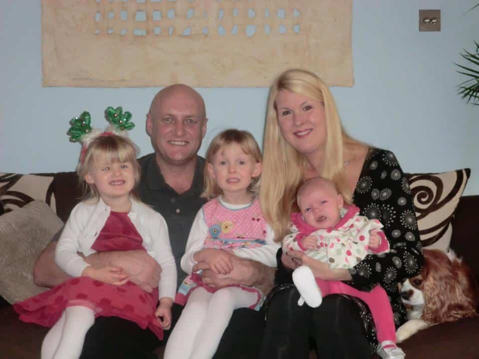 A family of five sits closely on a brown sofa: a bald man, blonde woman holding a baby, and two young girls, one wearing green festive headbands. A dog rests beside them. All are smiling, dressed in colourful clothes, with a patterned painting on the wall behind.