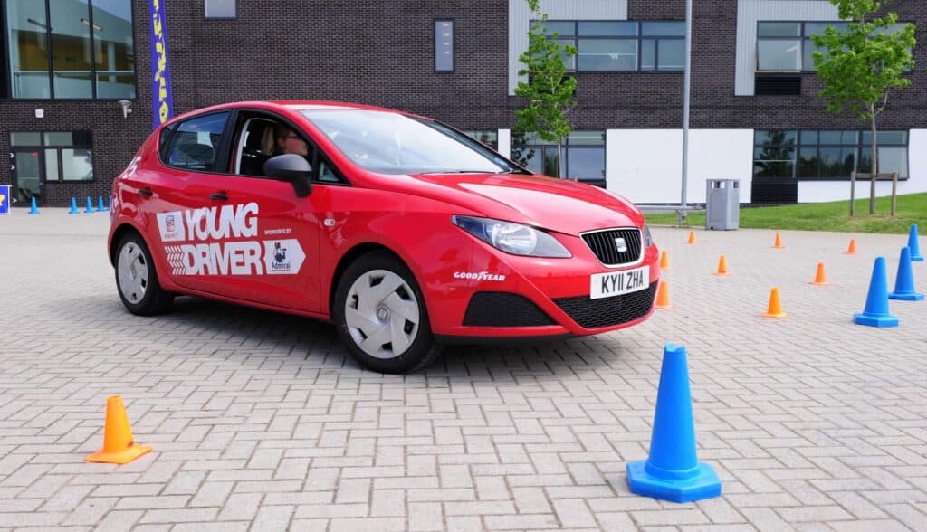 A red car with "Young Driver" branding is manoeuvring through an outdoor driving course marked by blue and orange traffic cones. A young person is visible in the driver's seat. The scene takes place on a paved area near a modern building with large windows.