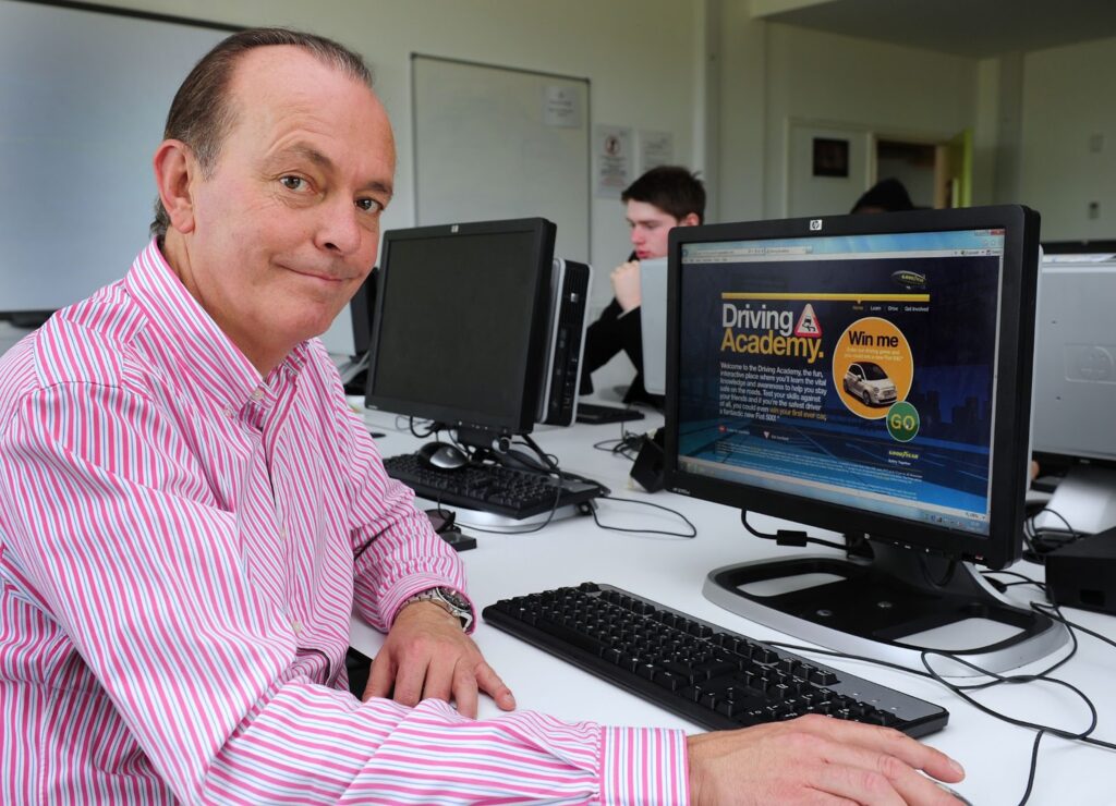 A middle-aged man in a pink striped shirt sits at a desk in a computer lab, smiling at the camera. He uses a desktop computer displaying a website for "Driving Academy." Other people are working at computers in the background.