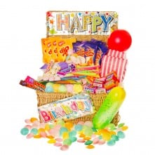 A wicker basket filled with colourful retro sweets, candy sticks, lollipops, fizzy sweets, a red balloon, a yellow balloon, and a striped popcorn bag. Signs reading "Happy" and "Birthday" are attached, creating a festive birthday display.