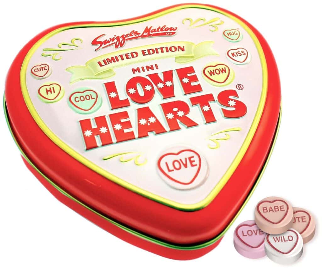 A red, heart-shaped tin labelled "Swizzels Matlow Limited Edition Mini Love Hearts" with pastel sweet heart designs. Beside the tin are four sweet hearts in pink and white with messages: "BABE," "LOVE," "WILD," and "SWEET.