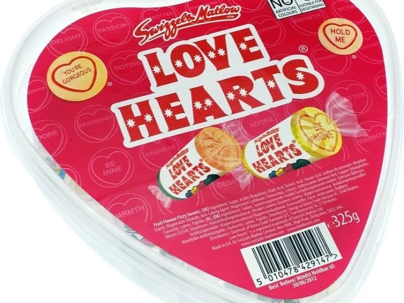 A heart-shaped container of Swizzels Matlow Love Hearts sweets. The red lid features the "Love Hearts" logo, phrases like "You're Gorgeous" and "Hold Me," and displays wrapped sweets inside. The container weighs 325g and has a barcode visible.