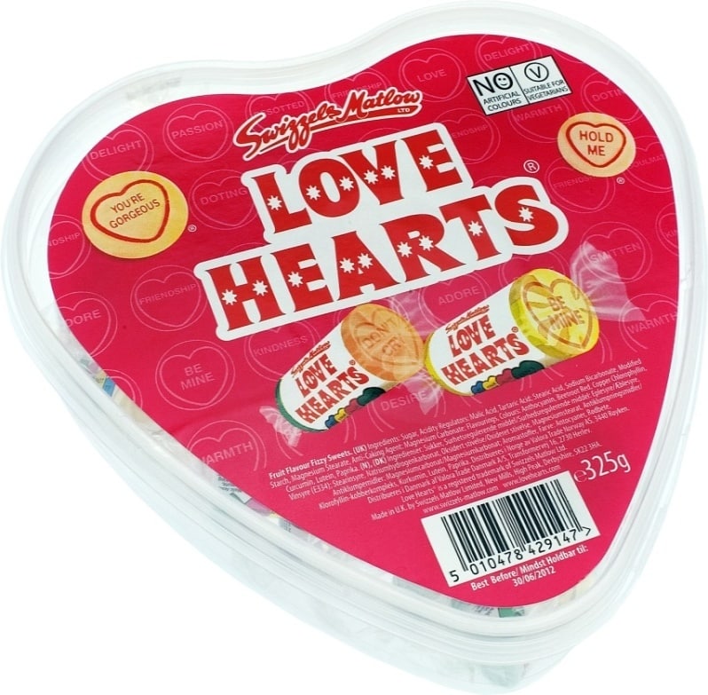 A heart-shaped container of Swizzels Matlow Love Hearts sweets. The red lid features the "Love Hearts" logo, phrases like "You're Gorgeous" and "Hold Me," and displays wrapped sweets inside. The container weighs 325g and has a barcode visible.