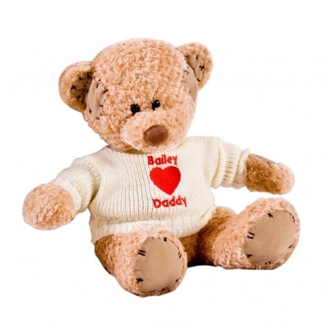 A soft, light brown teddy bear with black eyes and brown nose sits upright. It wears a white knitted jumper with red text reading "Bailey ♥ Daddy." The bear has darker brown fabric patches on its ears, paws, and body. The background is plain white.