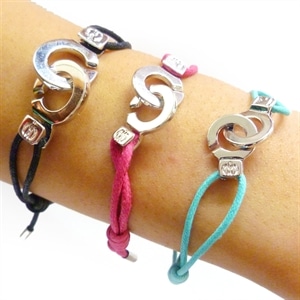 Three bracelets are worn on a forearm. Each has a silver interlocking "G" design centrepiece and a coloured cord: black, pink, and turquoise. The bracelets also feature small square metallic accents near the centrepiece.