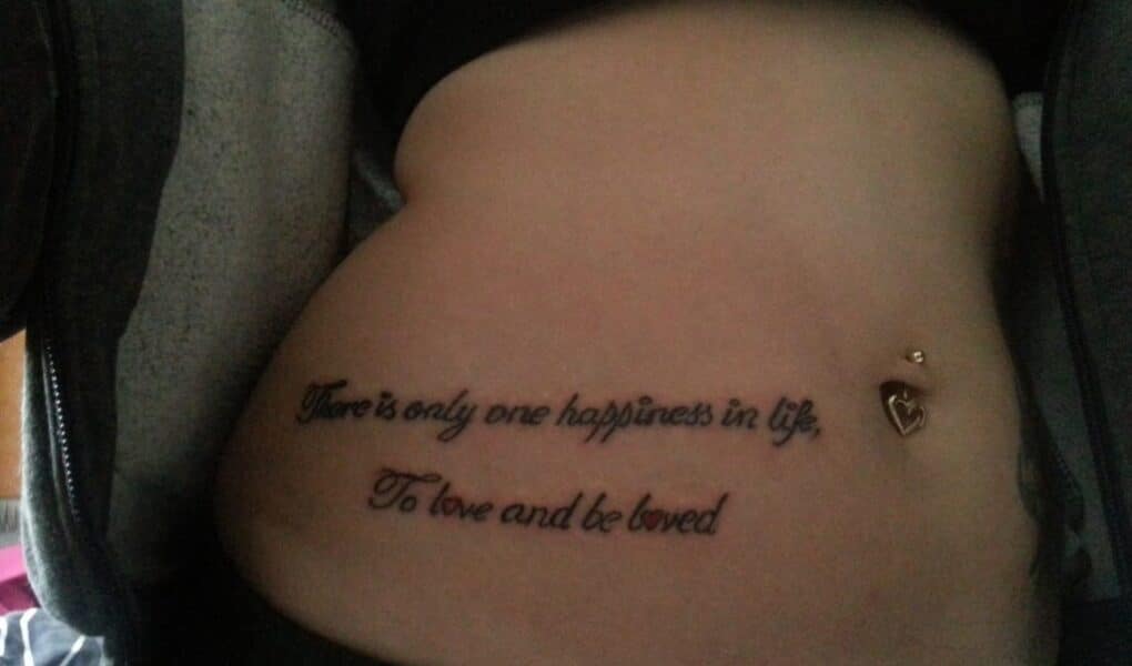 A tattoo on a person's lower abdomen reads, "There is only one happiness in life, To love and be loved." The person has a belly button piercing with a dangling heart charm and is wearing a partially unzipped hoodie.