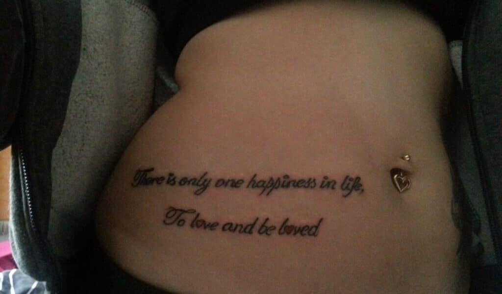 A tattoo on a person's lower abdomen reads, "There is only one happiness in life, To love and be loved." The person has a belly button piercing with a dangling heart charm and is wearing a partially unzipped hoodie.
