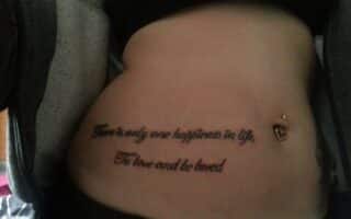 A tattoo on a person's lower abdomen reads, "There is only one happiness in life, To love and be loved." The person has a belly button piercing with a dangling heart charm and is wearing a partially unzipped hoodie.