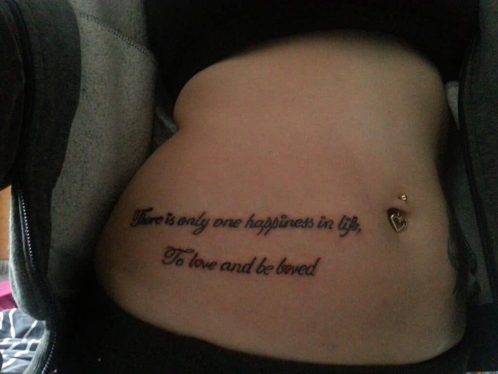 A close-up of a person's lower torso shows a tattoo in cursive font reading, "There is only one happiness in life, To love and be loved." The person has a gold heart-shaped belly button piercing and is partially dressed in dark clothing.