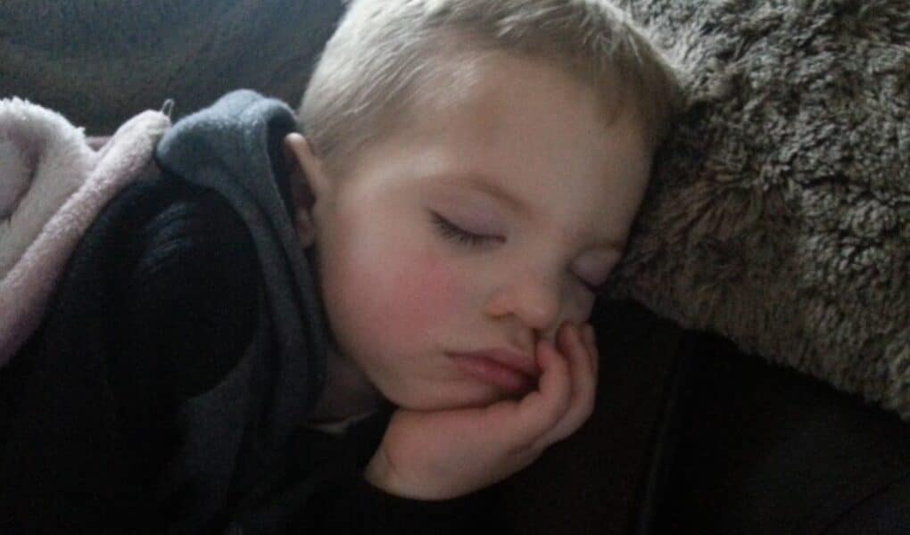 A young child with light brown hair sleeps peacefully, resting their head on a brown, fuzzy pillow. The child’s eyes are closed, and their hand is gently supporting their cheek. Soft lighting creates a calm and cosy atmosphere.
