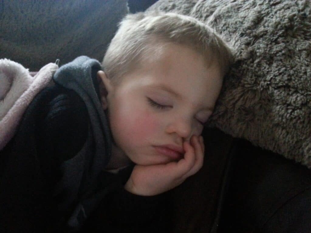 A young child with short blond hair is sleeping peacefully, resting their head on a fuzzy brown pillow. Their eyes are closed, and one hand supports their cheek. The child is covered partly with a pink blanket. The lighting is soft and natural.