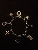 A silver charm bracelet with six different charms: a sunburst, a ring, a four-leaf clover, a round blue stone, a cross, a crescent moon, and a cluster of stones, displayed on a black background.