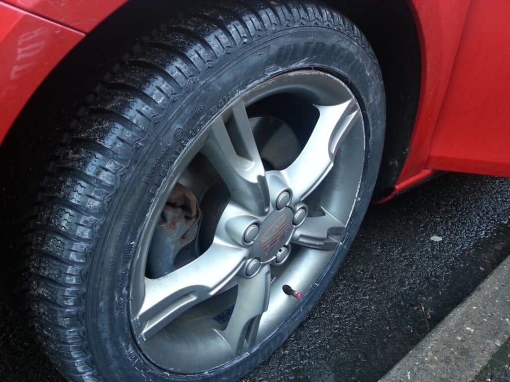 Close-up of a car’s front tyre and alloy wheel on a red vehicle. The tyre has visible tread and is slightly wet, suggesting recent rain. The car is parked on a damp tarmac surface near a concrete kerb. The wheel has five prominent spokes.