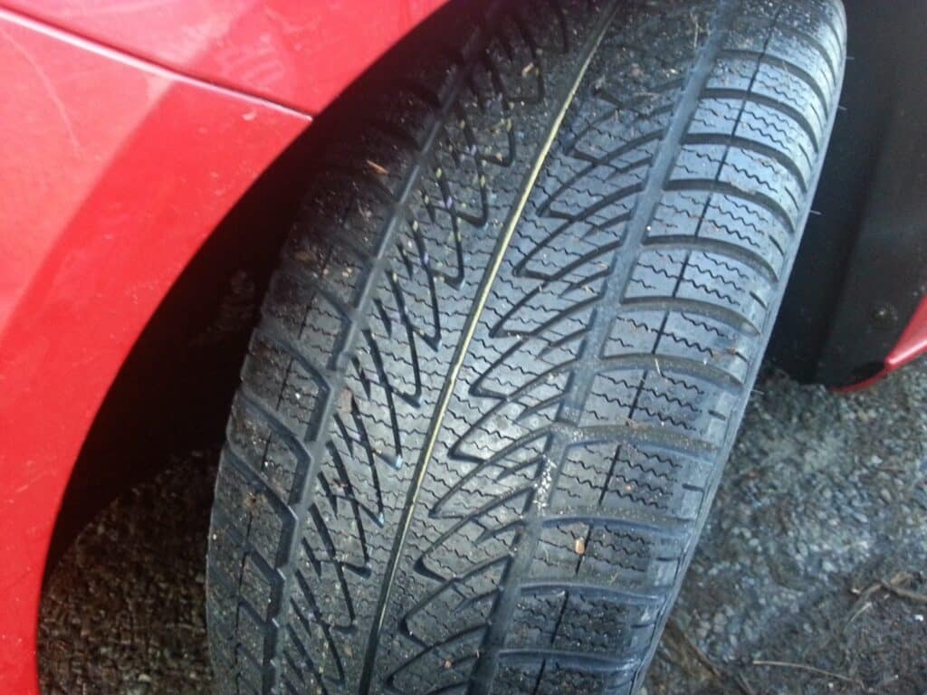 Close-up of a car tyre mounted on a red vehicle, showing detailed tread patterns with visible grooves and small stones embedded. The tyre appears to be in good condition, and the surrounding ground is rough with gravel and dirt.