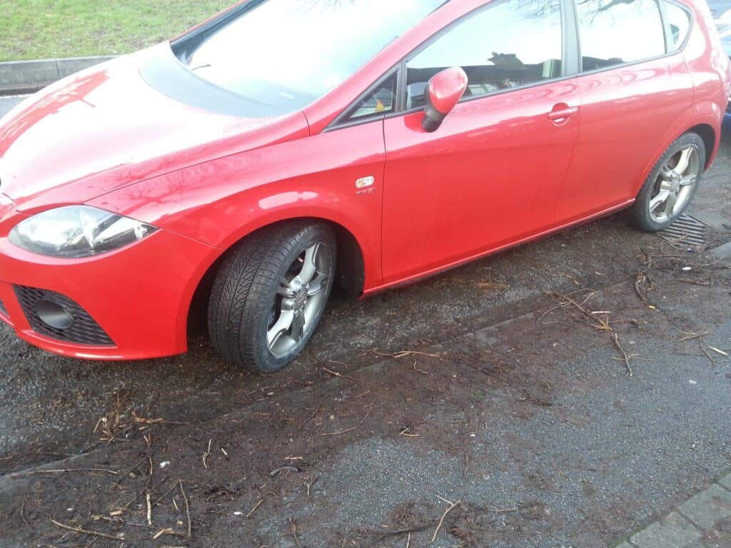 A red car is parked on the street with its front left wheel turned sharply outward, resting on the kerb. The car is slightly dirty. Twigs and debris are scattered on the wet pavement, and grass is visible in the background.