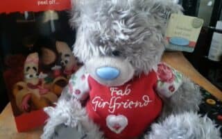 A grey teddy bear with a blue nose and floral-patterned arms sits upright. It wears a red shirt that says "Fab Girlfriend." Behind the bear is a red Valentine’s card and a coaster that reads “Handmade with Love.”.