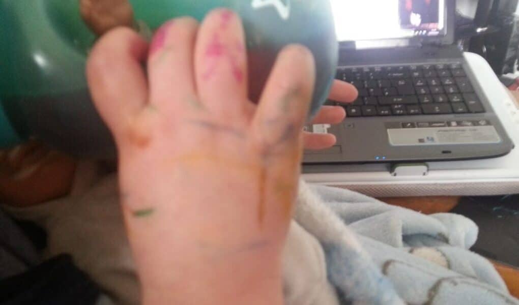 A baby’s hand covered in colourful felt-tip scribbles holds a green beaker. The baby is wrapped in a blue blanket. In the background, an open laptop sits on a cluttered desk.