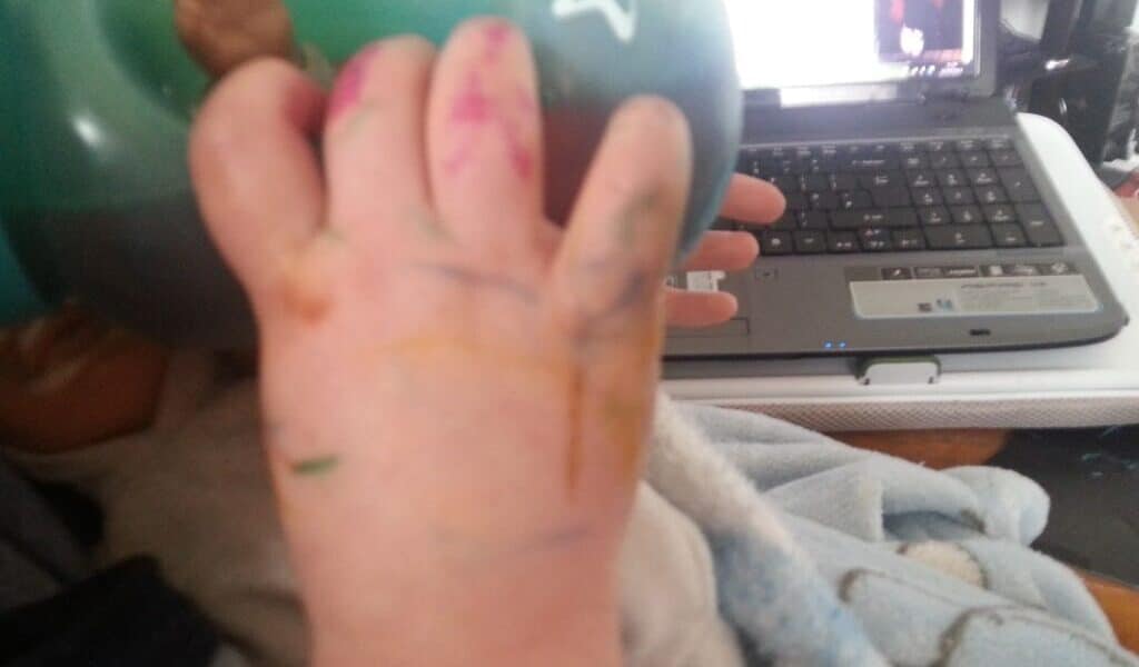 A baby’s hand covered in colourful felt-tip scribbles holds a green beaker. The baby is wrapped in a blue blanket. In the background, an open laptop sits on a cluttered desk.