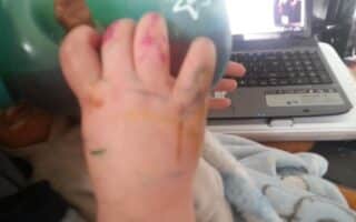 A baby’s hand covered in colourful felt-tip scribbles holds a green beaker. The baby is wrapped in a blue blanket. In the background, an open laptop sits on a cluttered desk.