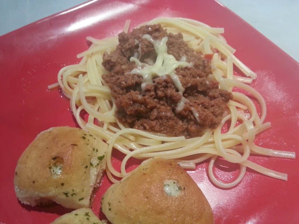 A red plate with a serving of spaghetti topped with minced beef meat sauce and a small amount of melted cheese in the centre. Two pieces of golden garlic bread with parsley are placed next to the pasta.