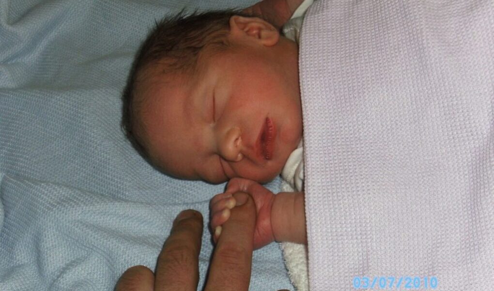 A newborn baby with dark hair sleeps on a light blue blanket, wrapped in a pale pink towel. The baby gently grasps an adult’s finger with its tiny hand. The date "03/07/2010" is visible in light blue text in the bottom right corner.