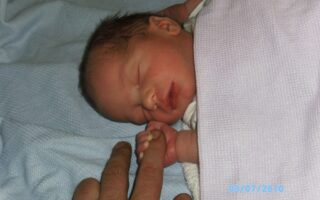 A newborn baby with dark hair sleeps on a light blue blanket, wrapped in a pale pink towel. The baby gently grasps an adult’s finger with its tiny hand. The date "03/07/2010" is visible in light blue text in the bottom right corner.