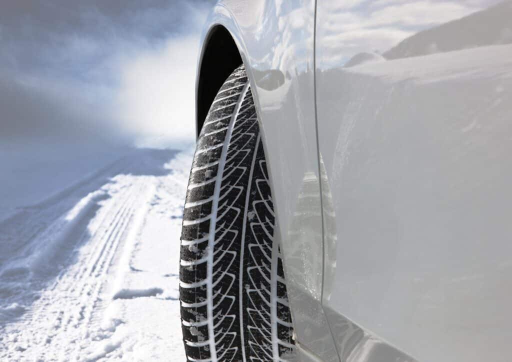 Close-up view of a car’s rear wheel fitted with a winter tyre, showing deep tread patterns. The car is parked or driving on a snow-covered road, with visible tyre tracks and snowy landscape in the background. The car’s body is white.