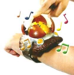 A child’s hand touches a large, colourful, egg-shaped toy worn on the wrist, decorated with orange and yellow details. Colourful musical notes float around, suggesting that the toy plays music or sounds. The background is plain white.