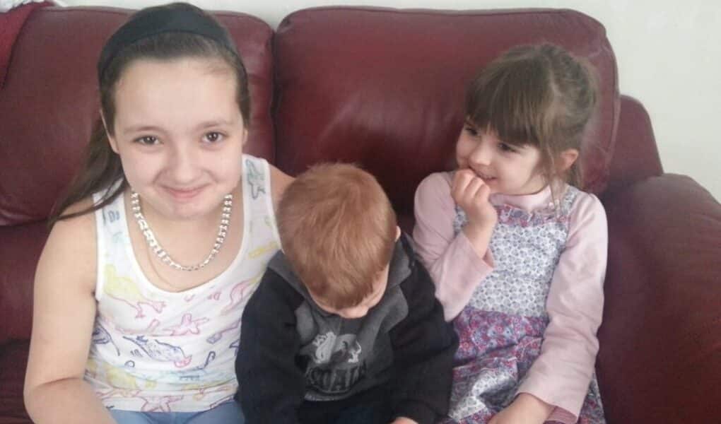 Three young children sit on a red leather sofa. The girl on the left smiles at the camera, wearing a white vest and bead necklace. The boy in the middle looks down, holding a blue bottle. The girl on the right, in a patterned dress, smiles with her hand by her mouth.