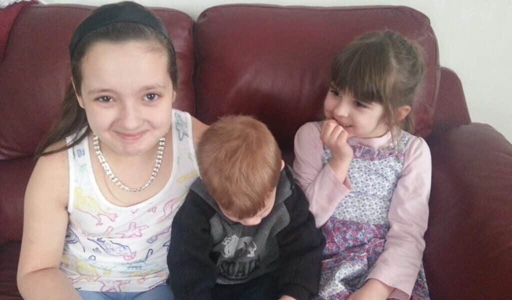 Three young children sit on a red leather sofa. The girl on the left smiles at the camera, wearing a white vest and bead necklace. The boy in the middle looks down, holding a blue bottle. The girl on the right, in a patterned dress, smiles with her hand by her mouth.