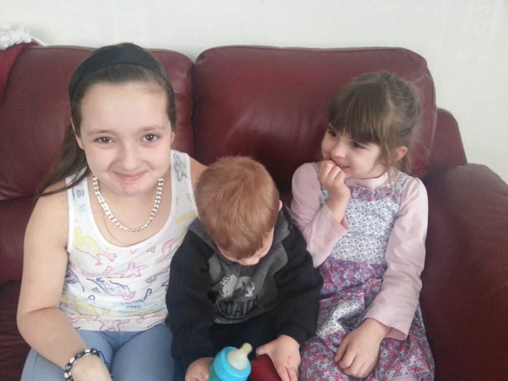 Three children sit on a red settee. On the left, a smiling girl with a headband and necklace looks at the camera. In the middle, a toddler with short ginger hair looks down, holding a blue bottle. On the right, a girl in a patterned dress smiles shyly.