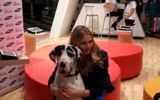 A blonde woman in a blue outfit kneels on a wooden floor, hugging a large black and white Great Dane. They are indoors at an event with red and orange round seats, promotional banners, and people in the background. Tablets are on the tables nearby.