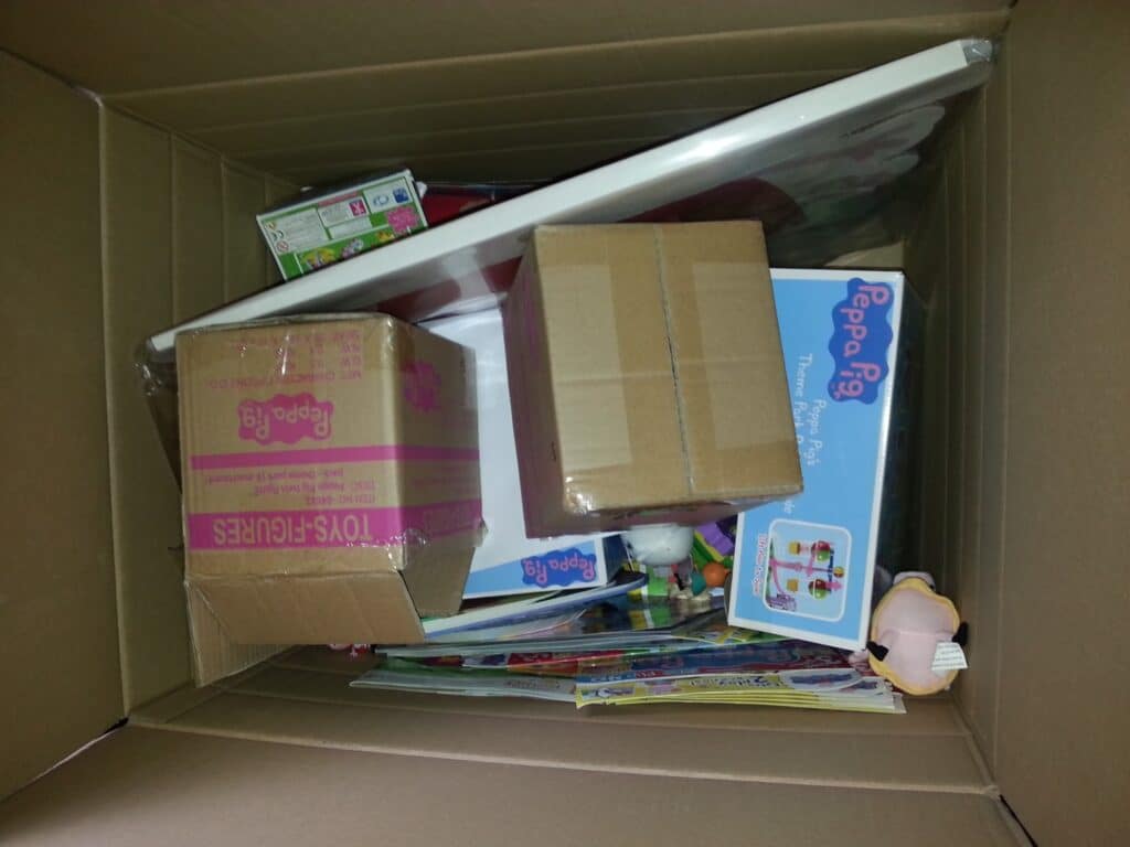 A large cardboard box filled with various toys and games, including boxed Peppa Pig items, a brown shipping box, a pink-striped box labelled "TOYS FIGURES," some colourful board games, and parts of soft toys visible around the sides.