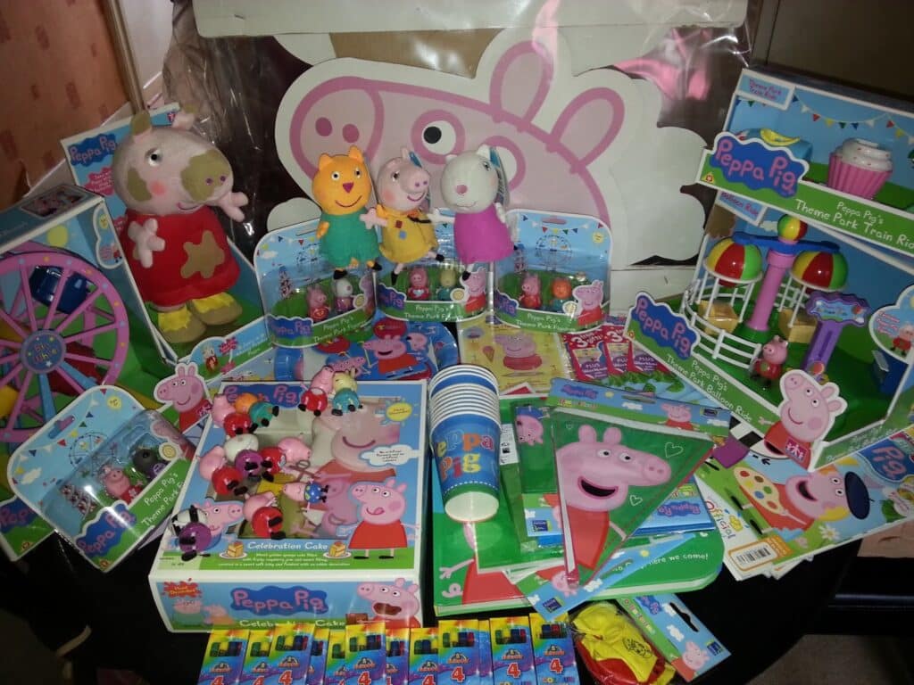 A table filled with various Peppa Pig-themed toys, books, party supplies, soft toys, figurines, and decorations. Most items feature Peppa Pig and her friends in bright, colourful packaging, creating a playful and festive atmosphere.