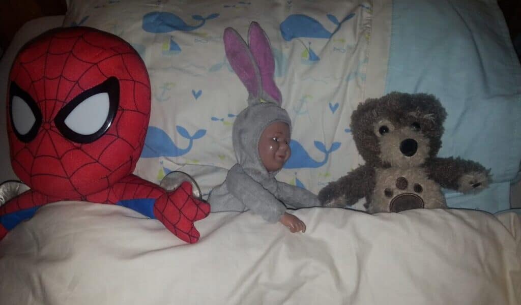 A plush Spiderman, a baby doll in a grey bunny suit, and a brown teddy bear are lying side by side under a white blanket on a bed with a pillow featuring a blue whale pattern.