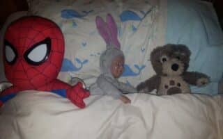A plush Spiderman, a baby doll in a grey bunny suit, and a brown teddy bear are lying side by side under a white blanket on a bed with a pillow featuring a blue whale pattern.