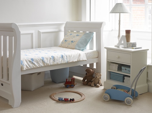 A cosy, sunlit child's bedroom with a white toddler bed, whale-patterned bedding, two stuffed bears, wooden toy train on a circular track, blue push toy, and a white bedside table with books, a lamp, and small decorative items, on a beige carpet.