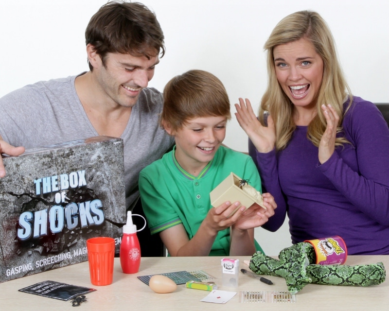 A smiling man, a boy in green holding an open box with a toy insect inside, and a surprised woman sit at a table with novelty prank items, including a rubber snake, a fake egg, trick cards, and “The Box of Shocks” game.