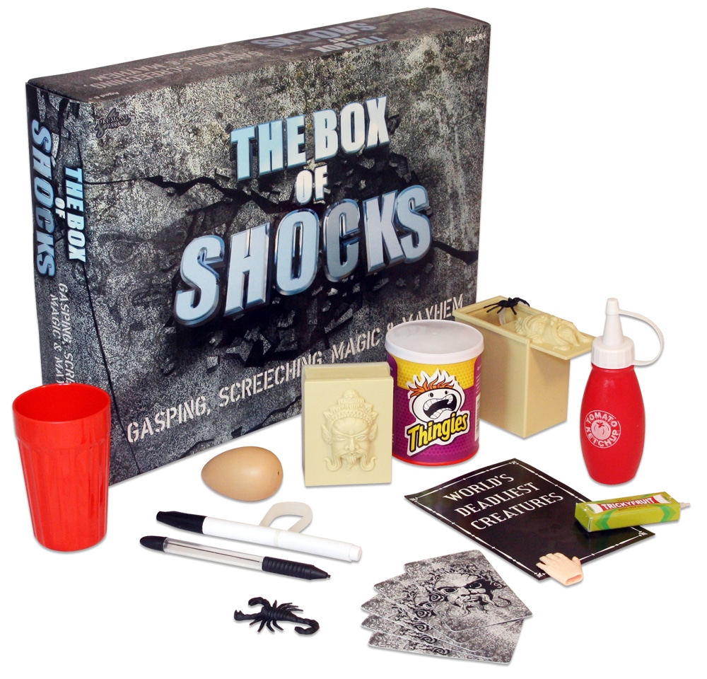 A magic prank kit titled "The Box of Shocks" includes a red cup, fake egg, novelty snack tin, creepy black spider, trick cards, two pens, a small bar of soap, a tomato ketchup bottle, prank boxes, and joke props, all arranged in front of the packaging.