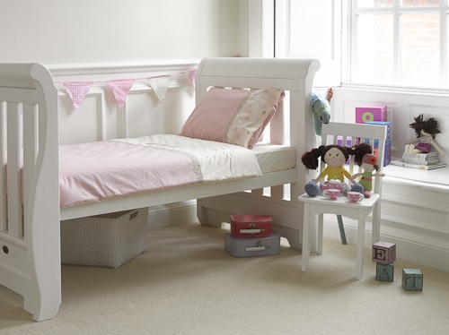 A cosy, light-filled child’s bedroom with a white sleigh bed dressed in pink and cream bedding. Toys, books, and pastel blocks sit on a small blue chair and white table. Pink bunting hangs on the wall above a window with white trim.