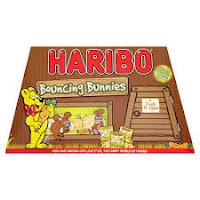 A Haribo box labelled "Bouncing Bunnies" features a yellow bear mascot with a red bowtie, cartoon bunnies in a window, a wooden door marked "Push to open," and images of gummy sweet bags. The background resembles wooden planks.