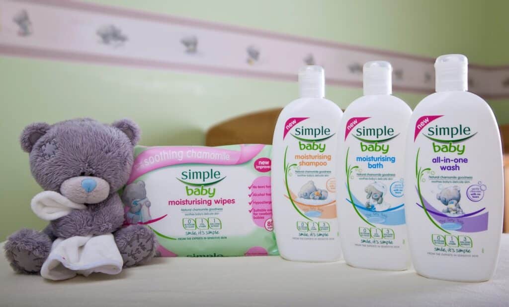A grey teddy bear holding a white cloth sits beside four Simple Baby products: moisturising wipes, moisturising shampoo, moisturising bath, and all-in-one wash, all featuring white bottles with coloured labels and a soft green background.