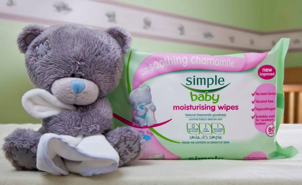 A grey teddy bear with a white cloth sits beside a packet of Simple Baby Moisturising Wipes. The wipes packet is green and white, labelled “soothing chamomile,” and contains 80 wipes. The background features a light green wall with a bear border.