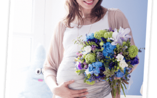 A pregnant woman in a white top and beige cardigan smiles whilst holding her belly and a large bouquet of purple, blue, and white flowers. A blue stuffed bunny sits in the background against a light blue wall near a sunlit window.