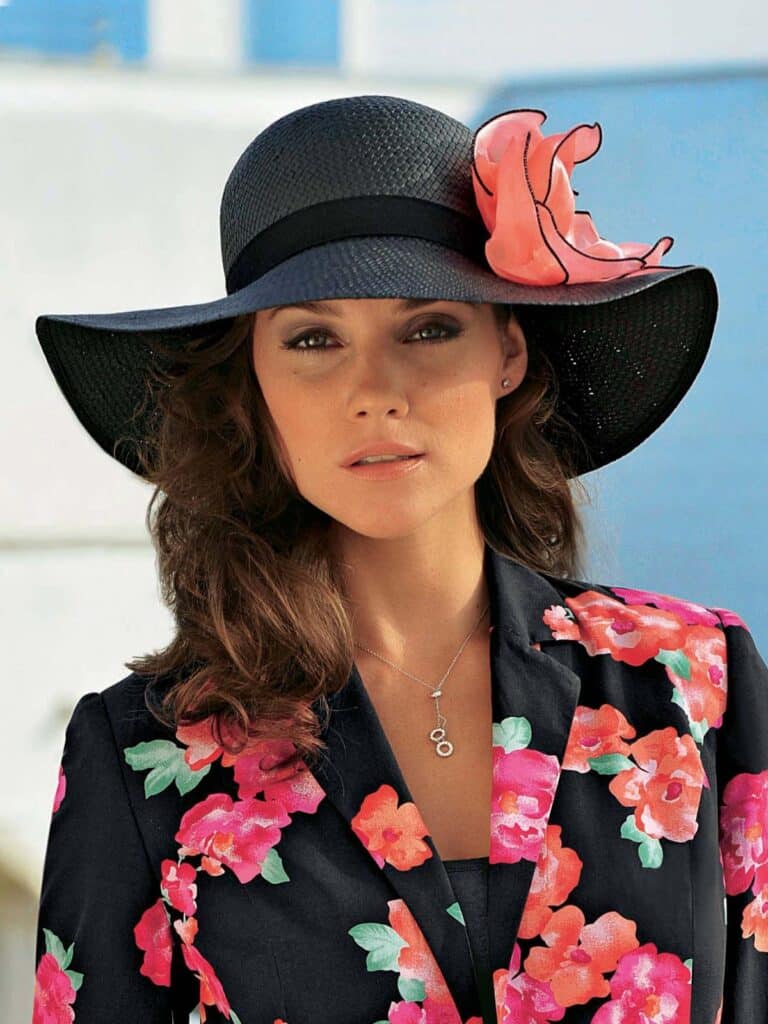 A woman with wavy brown hair wears a wide-brimmed black hat adorned with a large pink flower. She has a floral-patterned blazer with red and pink flowers over a black top and a delicate necklace. The background is softly focussed in light blue and white.