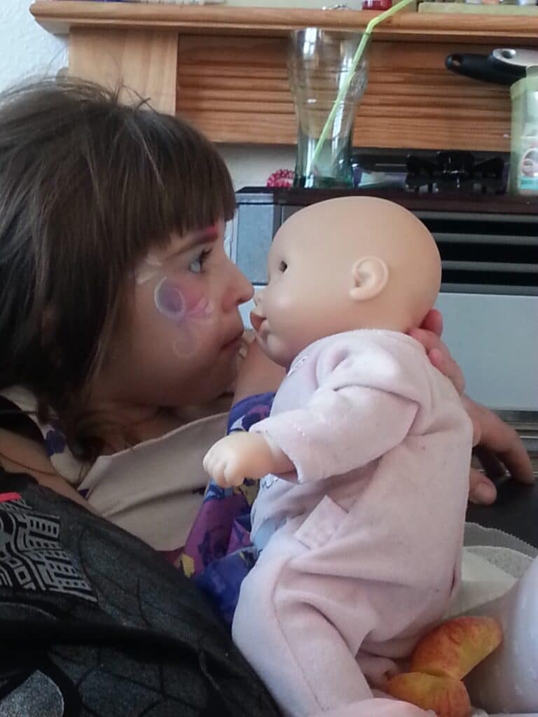 A young child with face paint gazes closely at a baby doll dressed in pink pyjamas. The child holds the doll gently, their noses almost touching. In the background, household items are visible on a shelf and a mantelpiece.