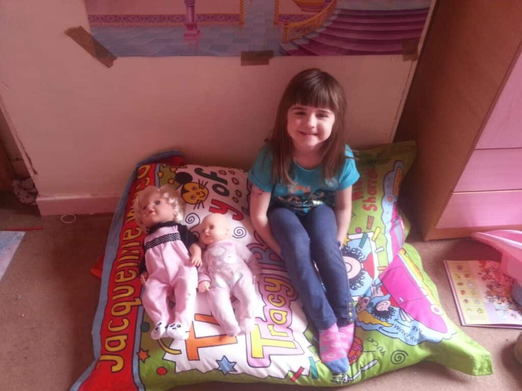 A young girl with brown hair and a fringe, wearing a blue T-shirt and jeans, sits cross-legged on a colourful Tracy Beaker-themed cushion. Two baby dolls, dressed in pink and white outfits, are beside her. The room has books, a chest of drawers, and a pink wall poster.